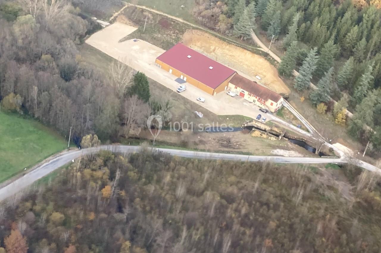 Vue aérienne d'un bâtiment avec un toit rouge entouré de forêt, d'une route et d'un petit cours d'eau.