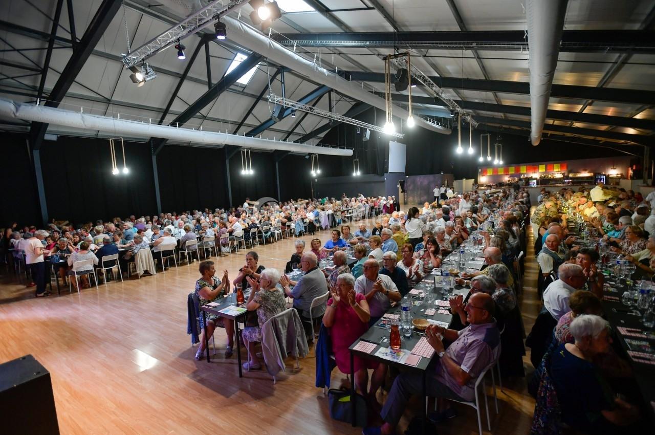 Salle comble avec de nombreuses personnes assises à des tables, participant à un événement convivial dans un grand espace…