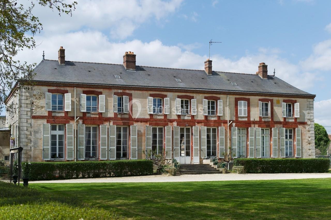Façade d'un grand bâtiment en pierre et briques rouges avec de nombreuses fenêtres, entouré d'un jardin.
