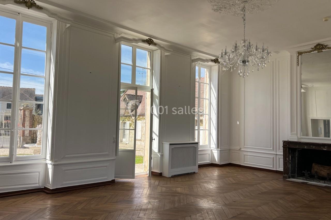 Salle lumineuse avec parquet en bois, grandes fenêtres donnant sur un jardin, cheminée et lustre décoratif.