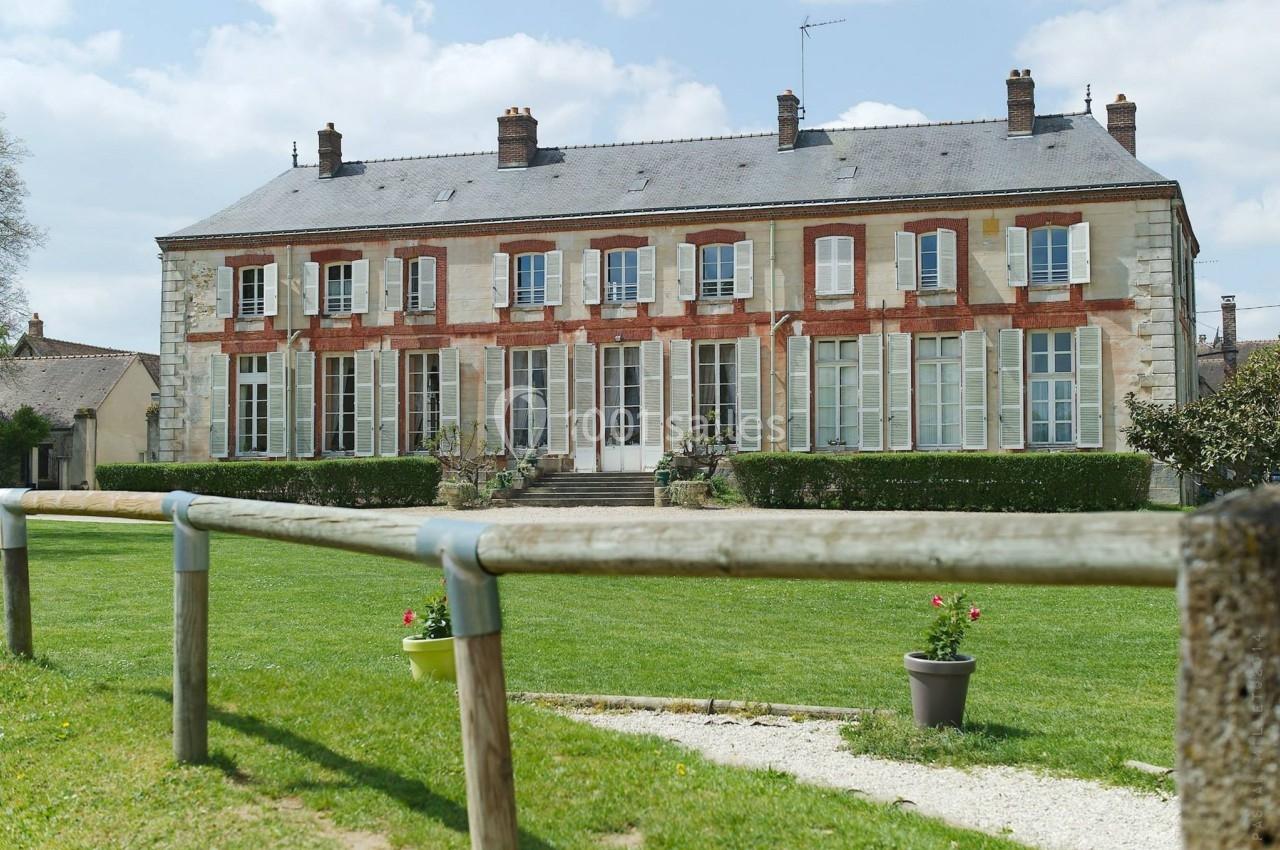 Grande maison en pierre et briques avec volets blancs, entourée d'une pelouse et de quelques pots de fleurs.