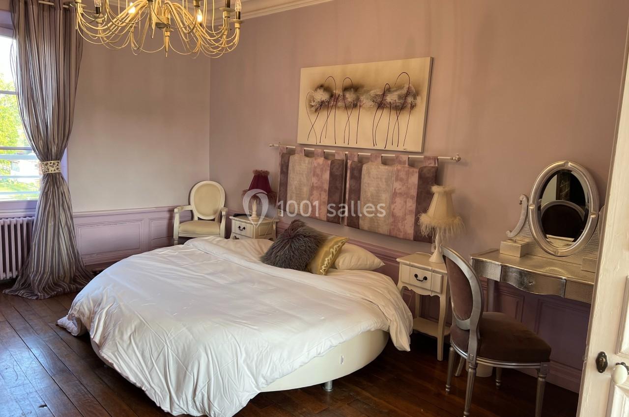 Chambre élégante avec lit rond, mobilier classique, murs mauves et lustre doré, éclairée par une fenêtre.