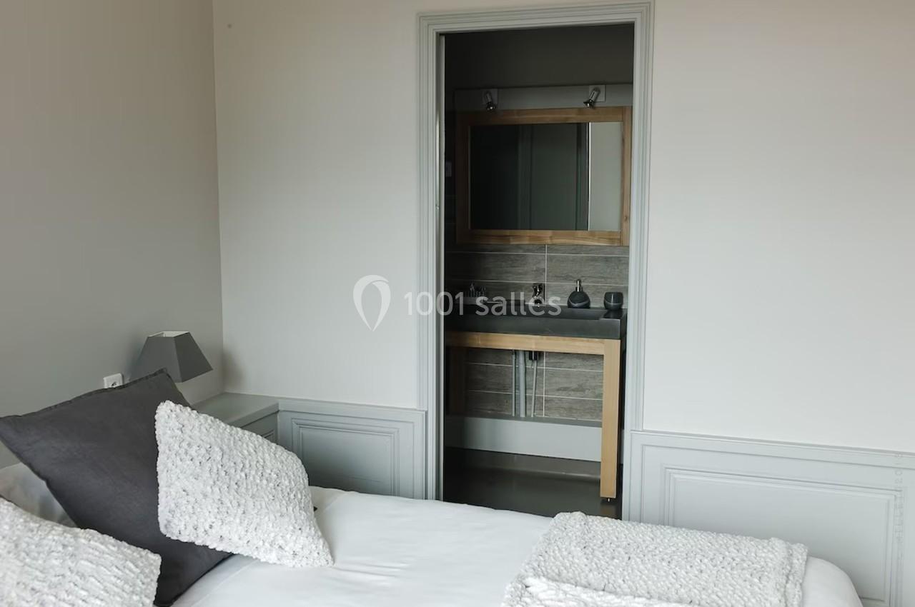 Vue d'une chambre avec un lit blanc et des coussins gris, donnant sur une salle de bain avec un miroir et un lavabo.