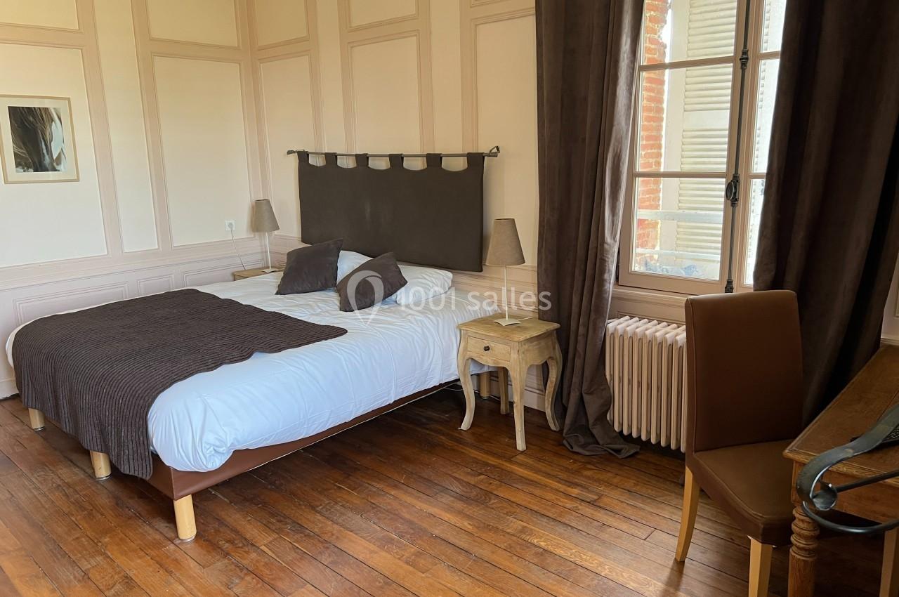 Chambre lumineuse avec lit double, parquet en bois, bureau, chaise, fenêtre avec rideaux et table de chevet.