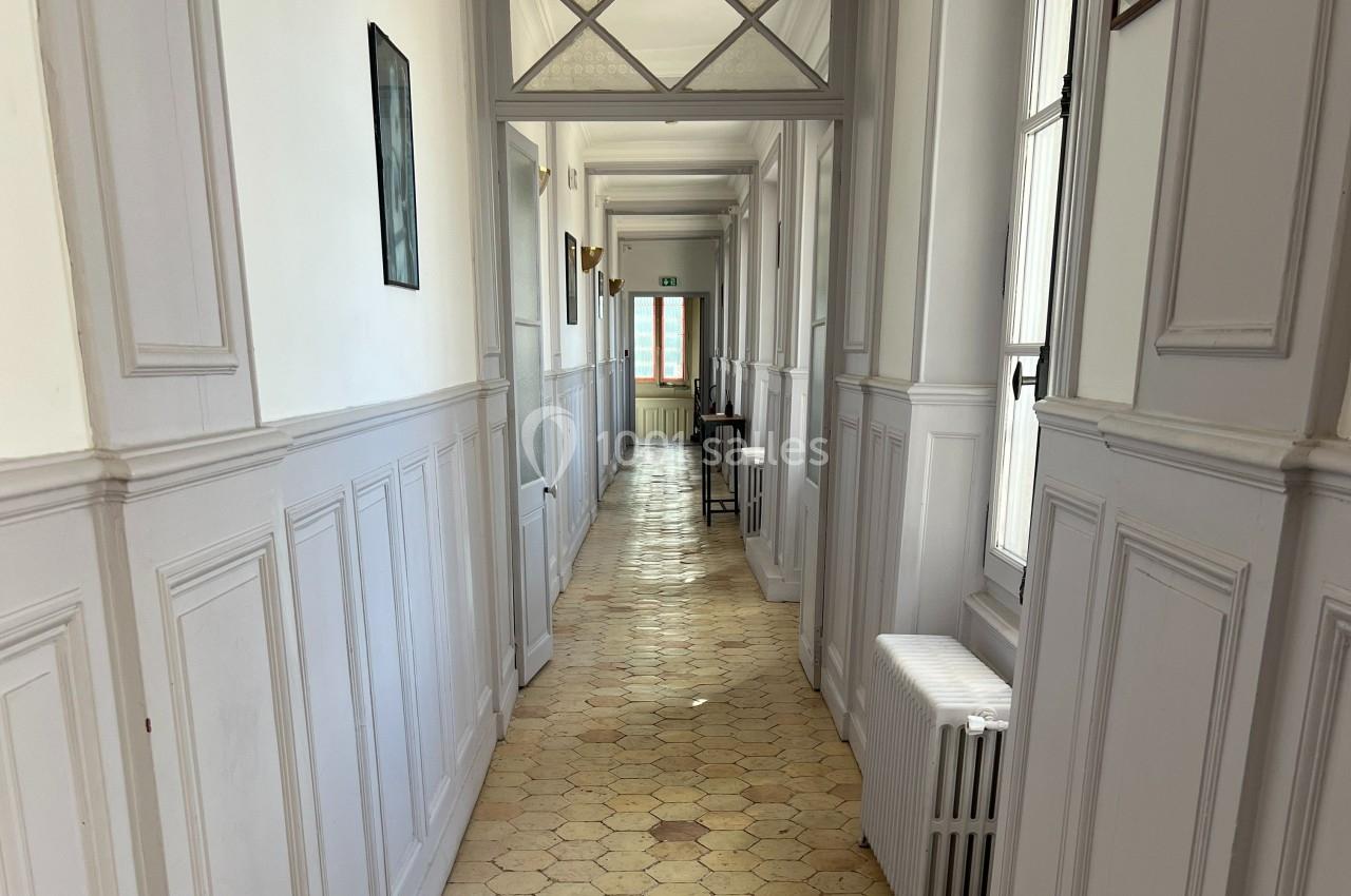 Couloir lumineux avec murs blancs, sol en carrelage beige, fenêtres à droite et porte au fond.
