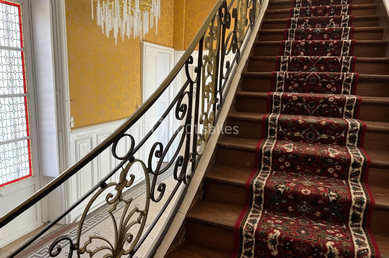 Escalier en bois avec rampe en fer forgé et tapis rouge à motifs, dans un intérieur aux murs jaunes et lustre en cristal.