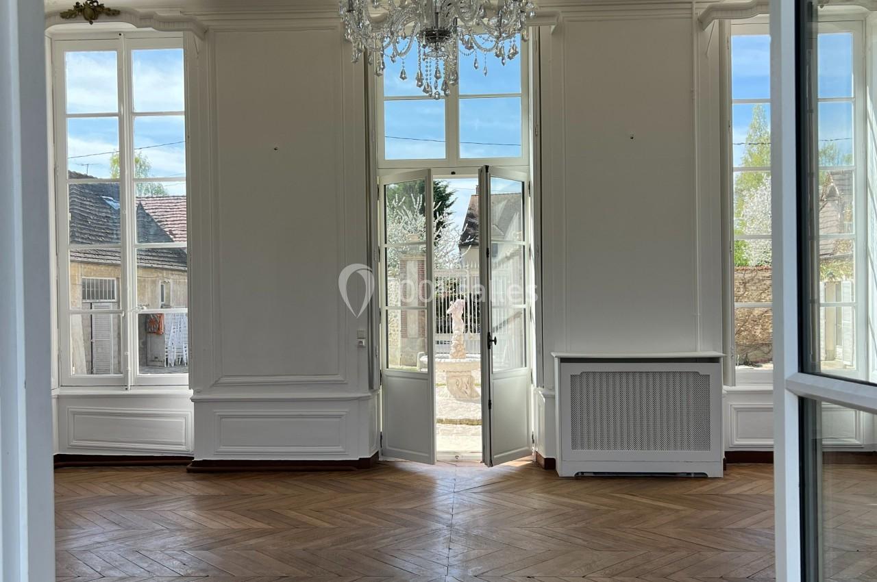 Pièce lumineuse avec parquet en bois, grandes fenêtres et porte-fenêtre donnant sur un extérieur.