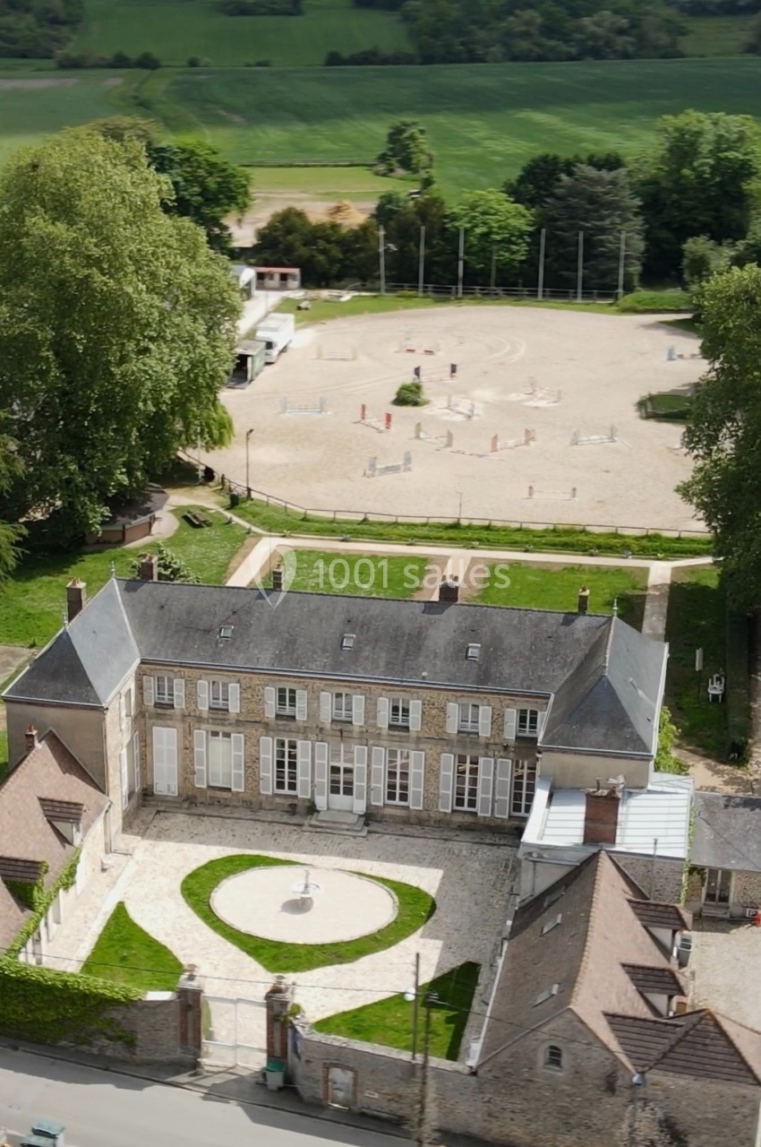 Vue aérienne d'un manoir en pierre entouré de jardins, avec une grande cour et un espace équestre en arrière-plan.