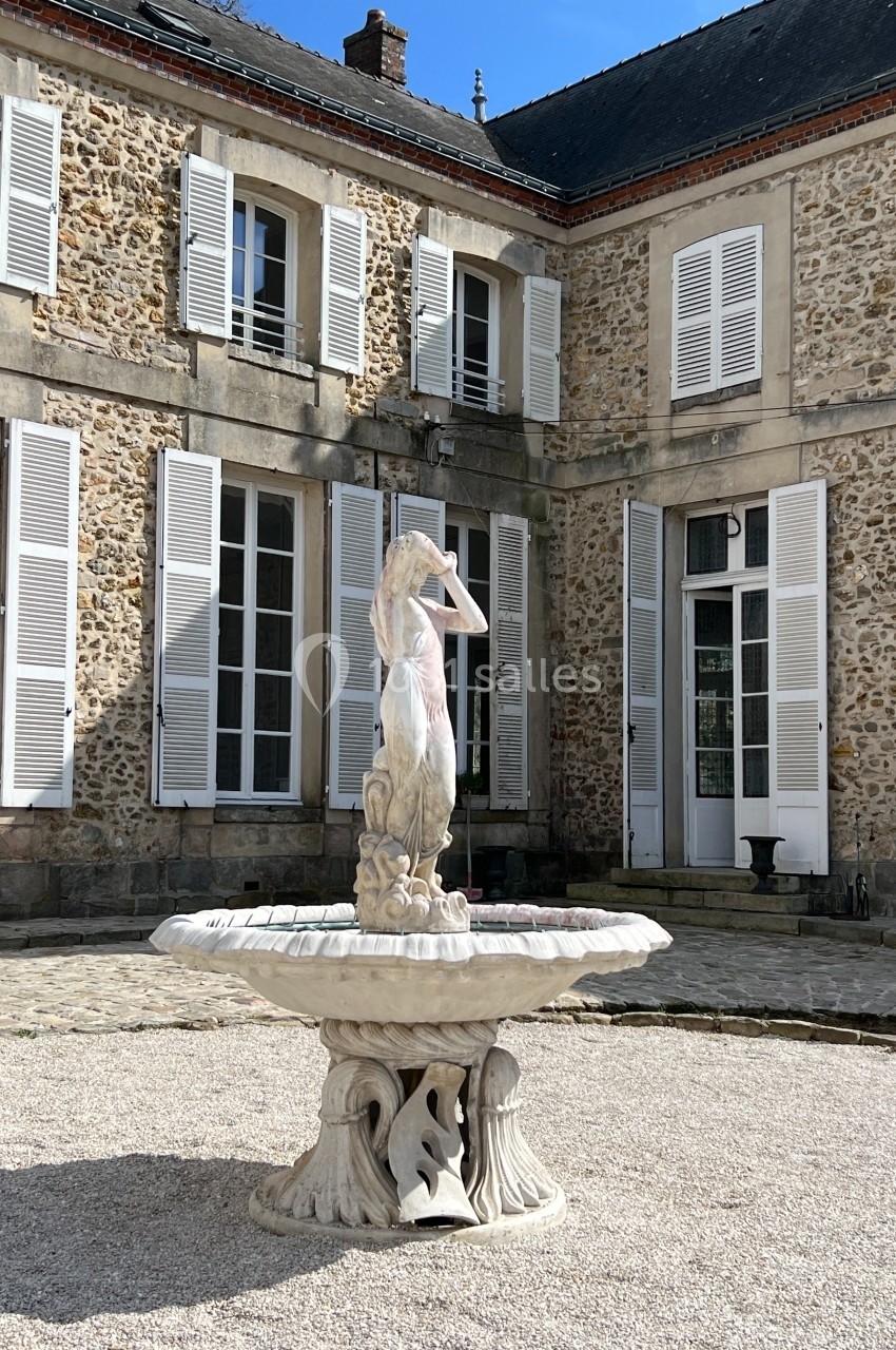 Fontaine décorative en pierre avec une statue centrale, située dans la cour pavée d'une maison en pierre aux volets blancs.