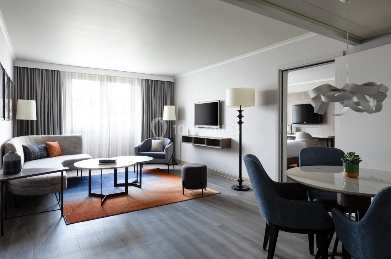 Salon moderne avec canapé, table basse, télé murale, coin repas et grande fenêtre laissant entrer la lumière naturelle.