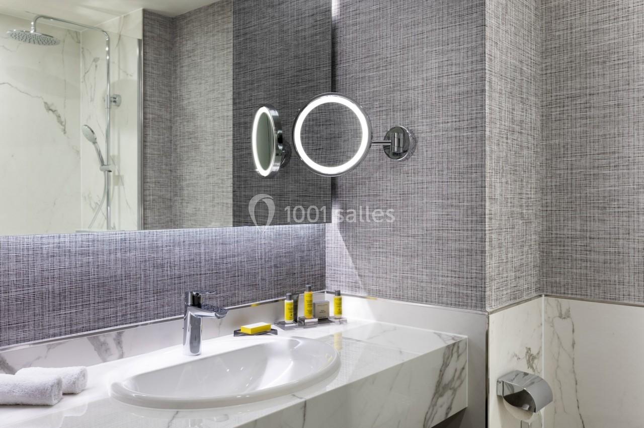 Salle de bain moderne avec lavabo en marbre, miroir éclairé, accessoires de toilette et douche en arrière-plan.