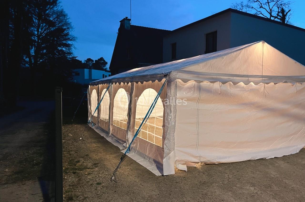 Tente blanche éclairée de l'intérieur, installée à l'extérieur près d'une maison au crépuscule.