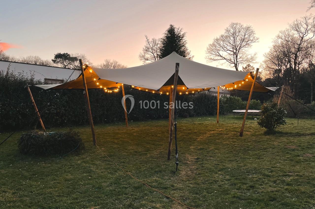 Tente beige éclairée par des guirlandes lumineuses, installée dans un jardin au coucher du soleil.
