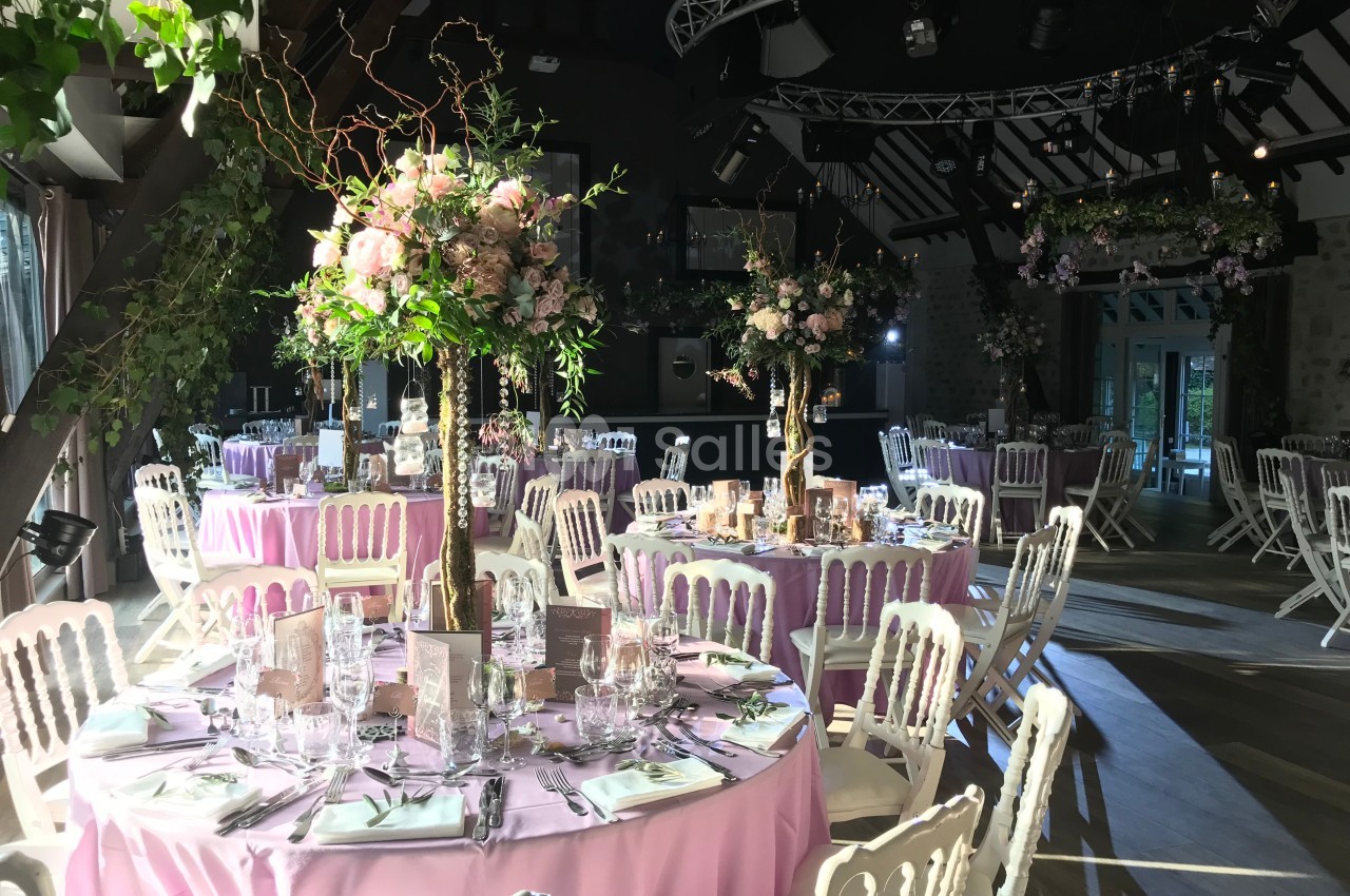 Salle de réception décorée avec des tables rondes, nappes roses, chaises blanches et compositions florales élégantes.