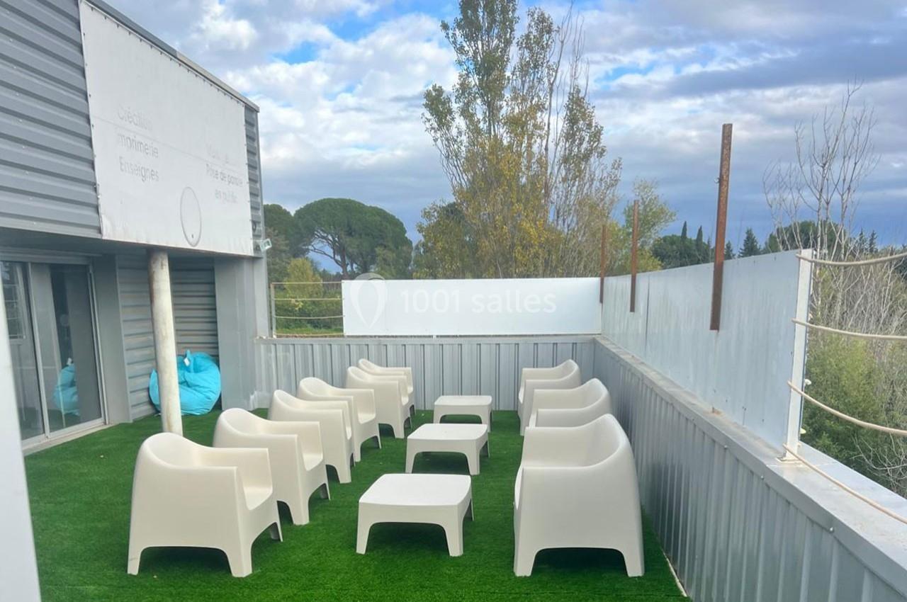 Terrasse extérieure avec fauteuils et tables blanches sur gazon synthétique, entourée de panneaux métalliques.