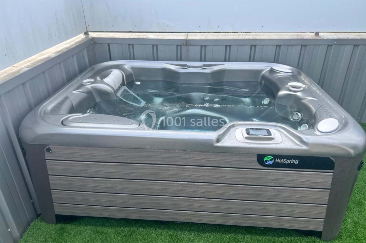 Bain à remous extérieur gris sur une surface en gazon synthétique, entouré de panneaux métalliques.