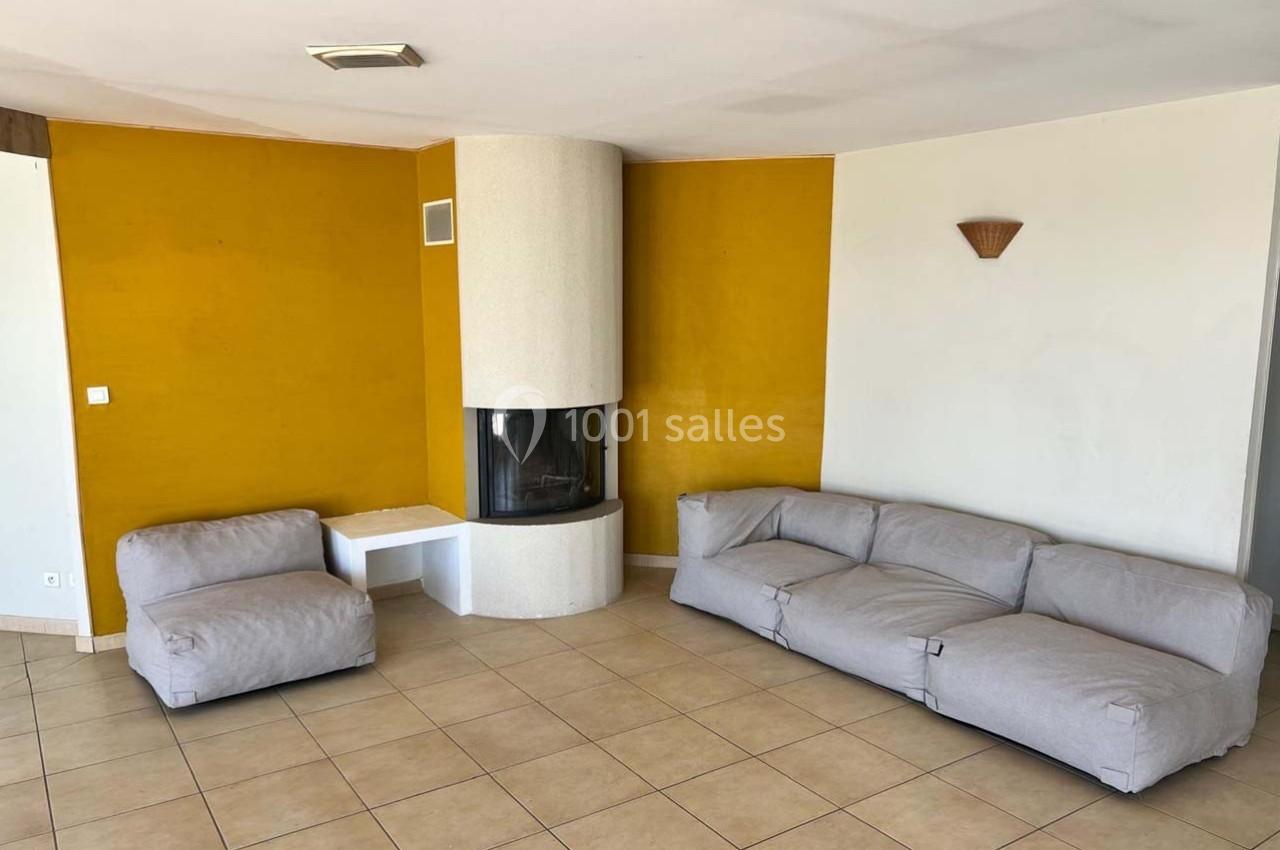 Salon avec murs jaunes, cheminée d'angle blanche, canapé gris en L et sol en carrelage beige.