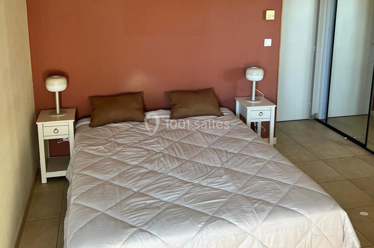 Lit double avec une couette blanche, deux oreillers bruns, tables de chevet blanches et lampes, mur rouge en arrière-plan.