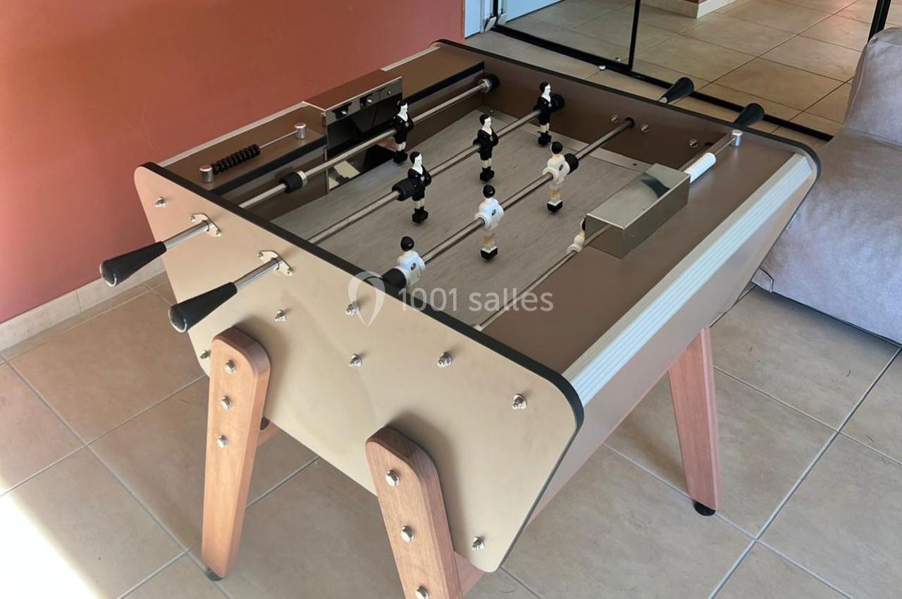 Table de baby-foot beige avec des joueurs noirs et blancs, posée sur un sol carrelé devant un mur rouge.