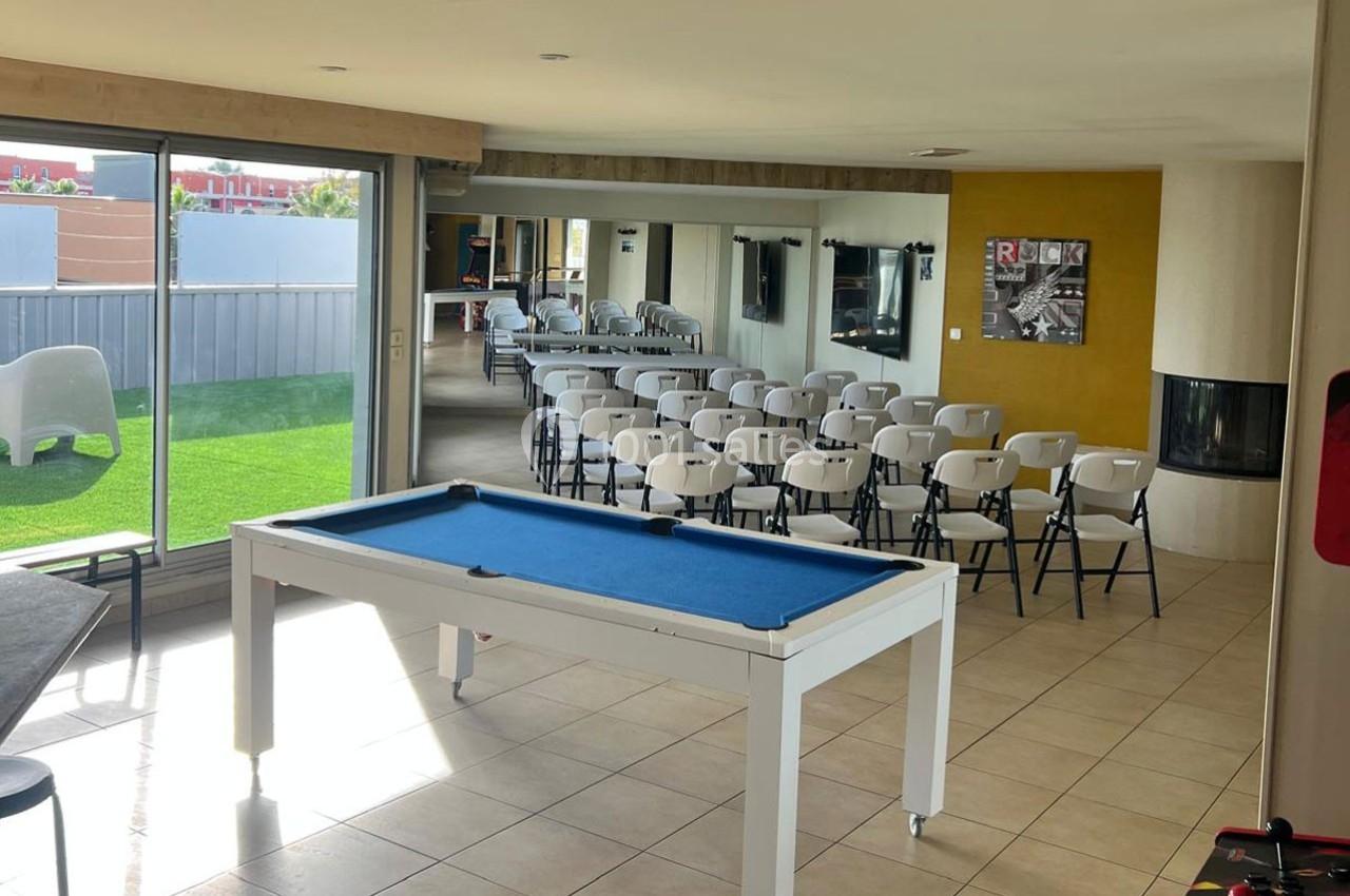 Salle lumineuse avec un billard, des chaises alignées pour une réunion, et une baie vitrée donnant sur un espace extérieur.