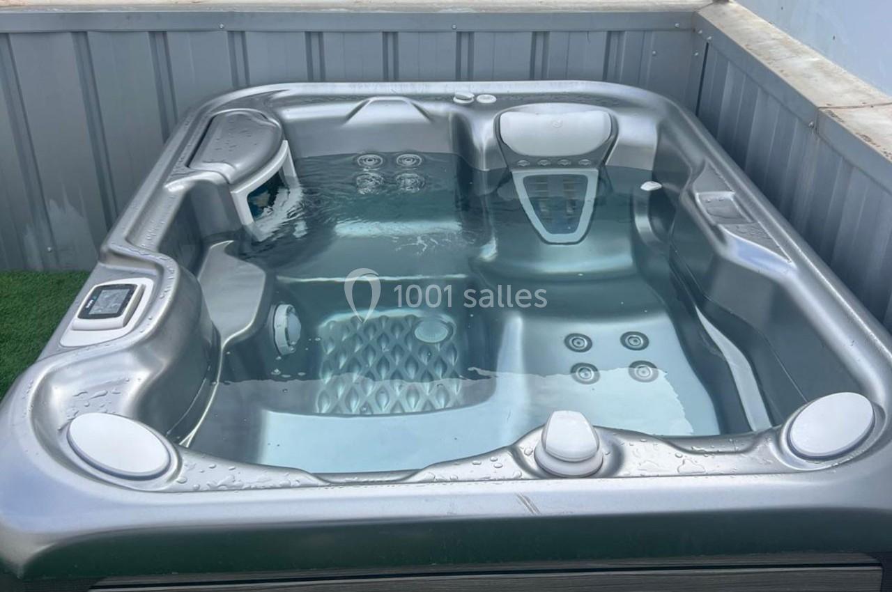 Spa extérieur gris rempli d'eau, installé sur une surface en gazon synthétique et entouré de parois métalliques.