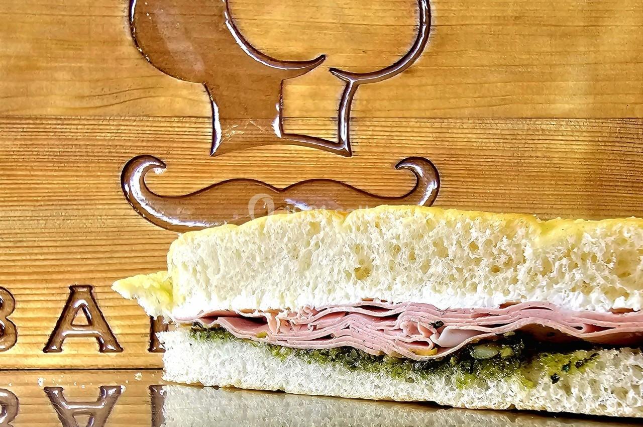 Sandwich garni de jambon et de pesto, présenté devant une planche en bois gravée d'un motif de chef.