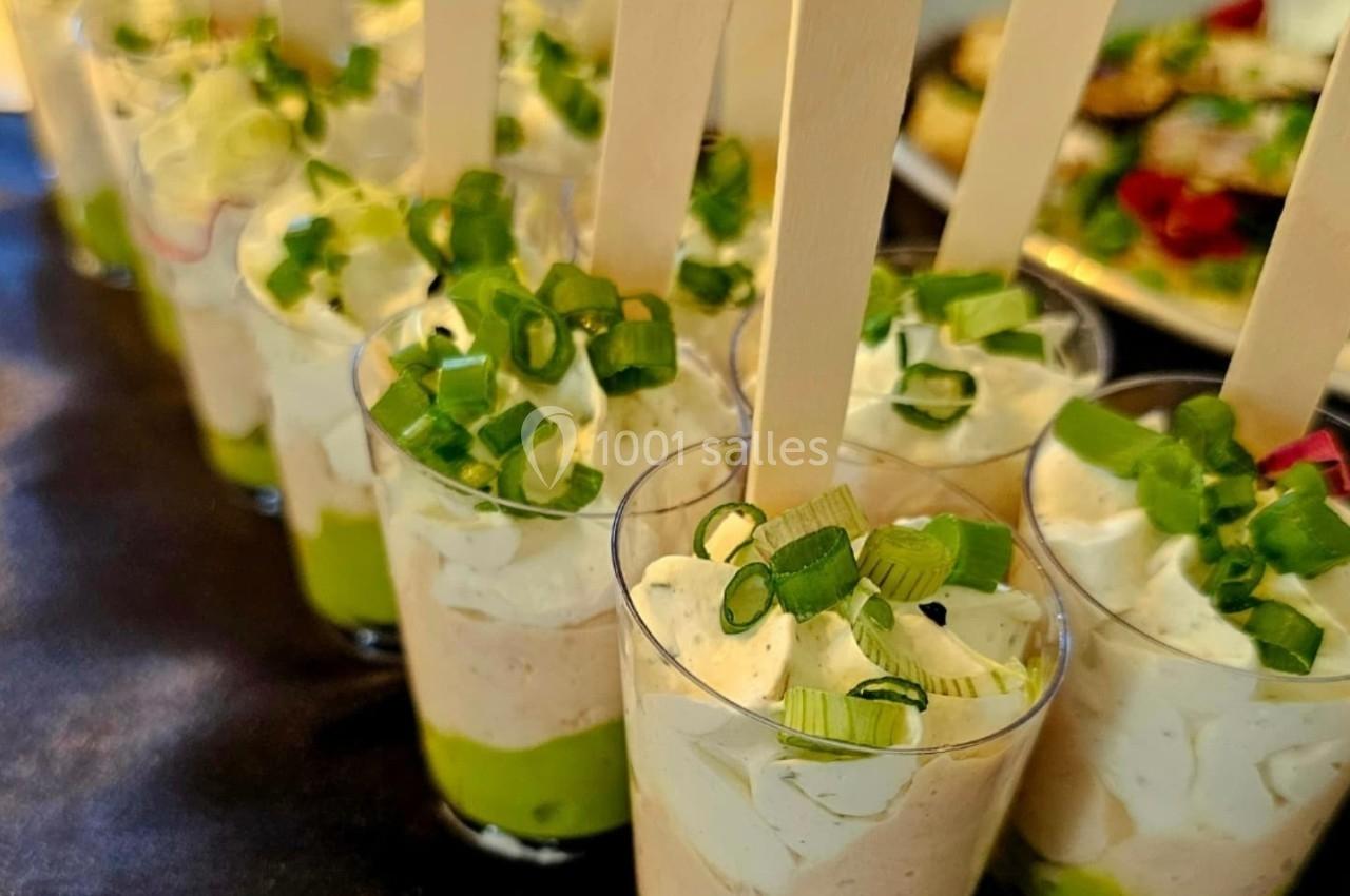 Verrines garnies de mousse verte, crème blanche, ciboulette et bâtonnets en bois, présentées sur une table.