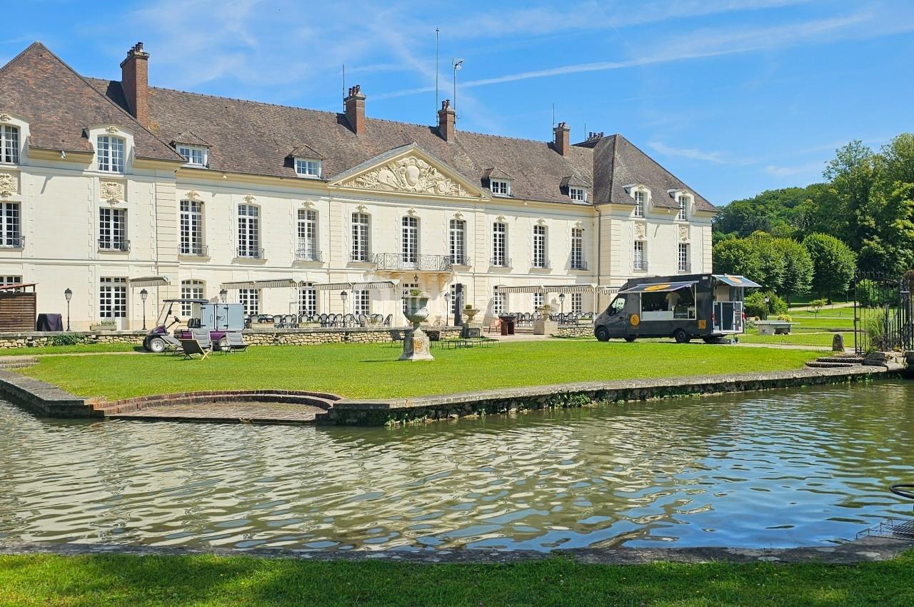 Façade d'un château bordant un étang, avec un food truck et des tables installées sur la pelouse.