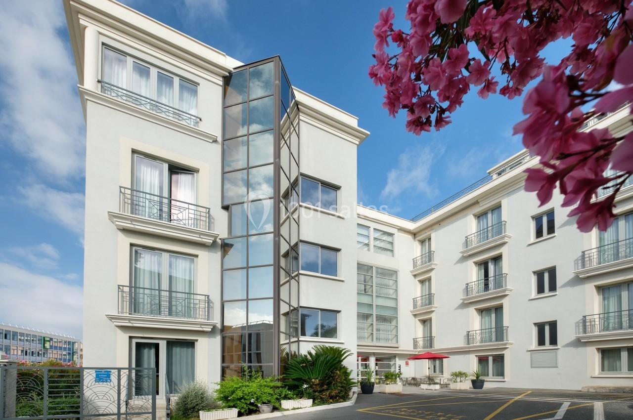 Façade d'un immeuble moderne avec grandes fenêtres, balcons, végétation et ciel bleu, vue partielle d'un arbre fleuri.