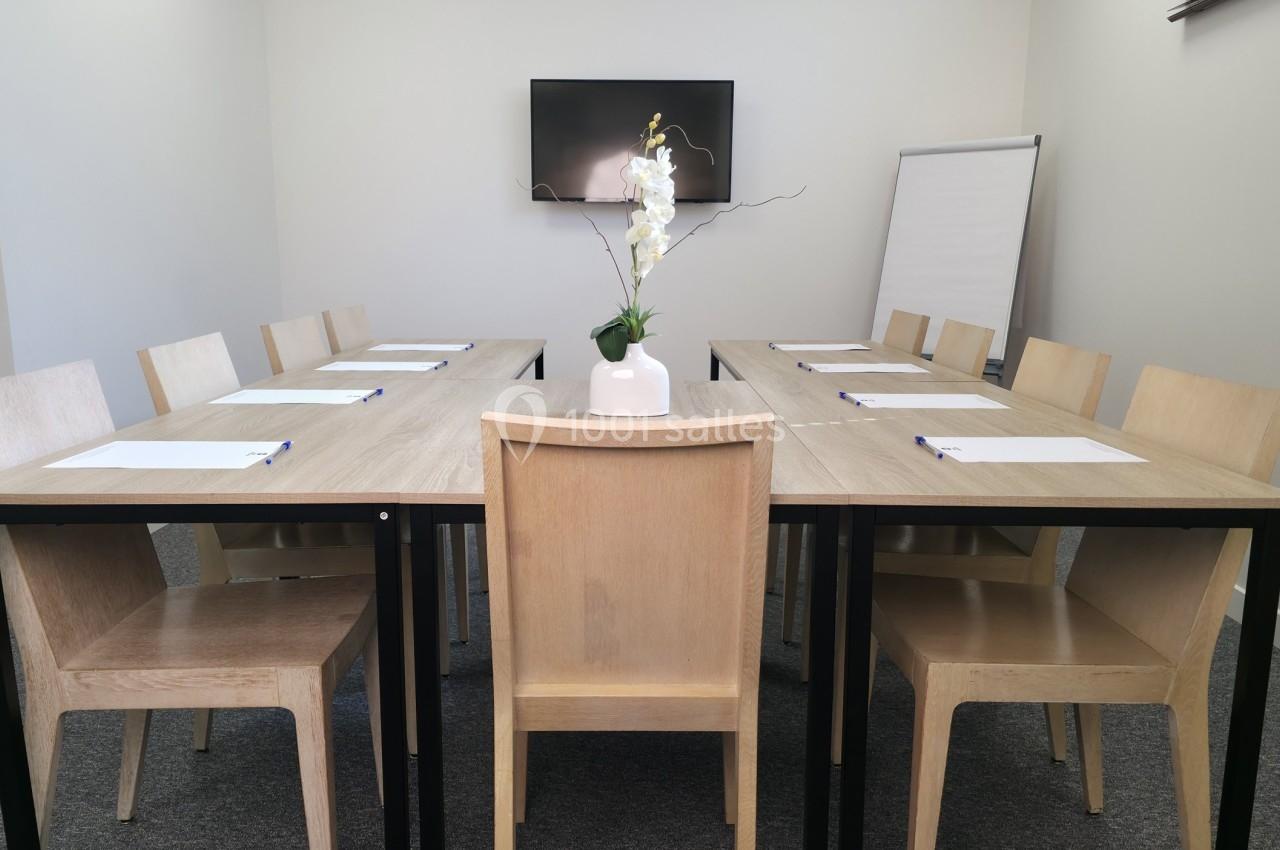 Salle de réunion avec table en bois, chaises assorties, orchidée au centre, écran mural et tableau de présentation.
