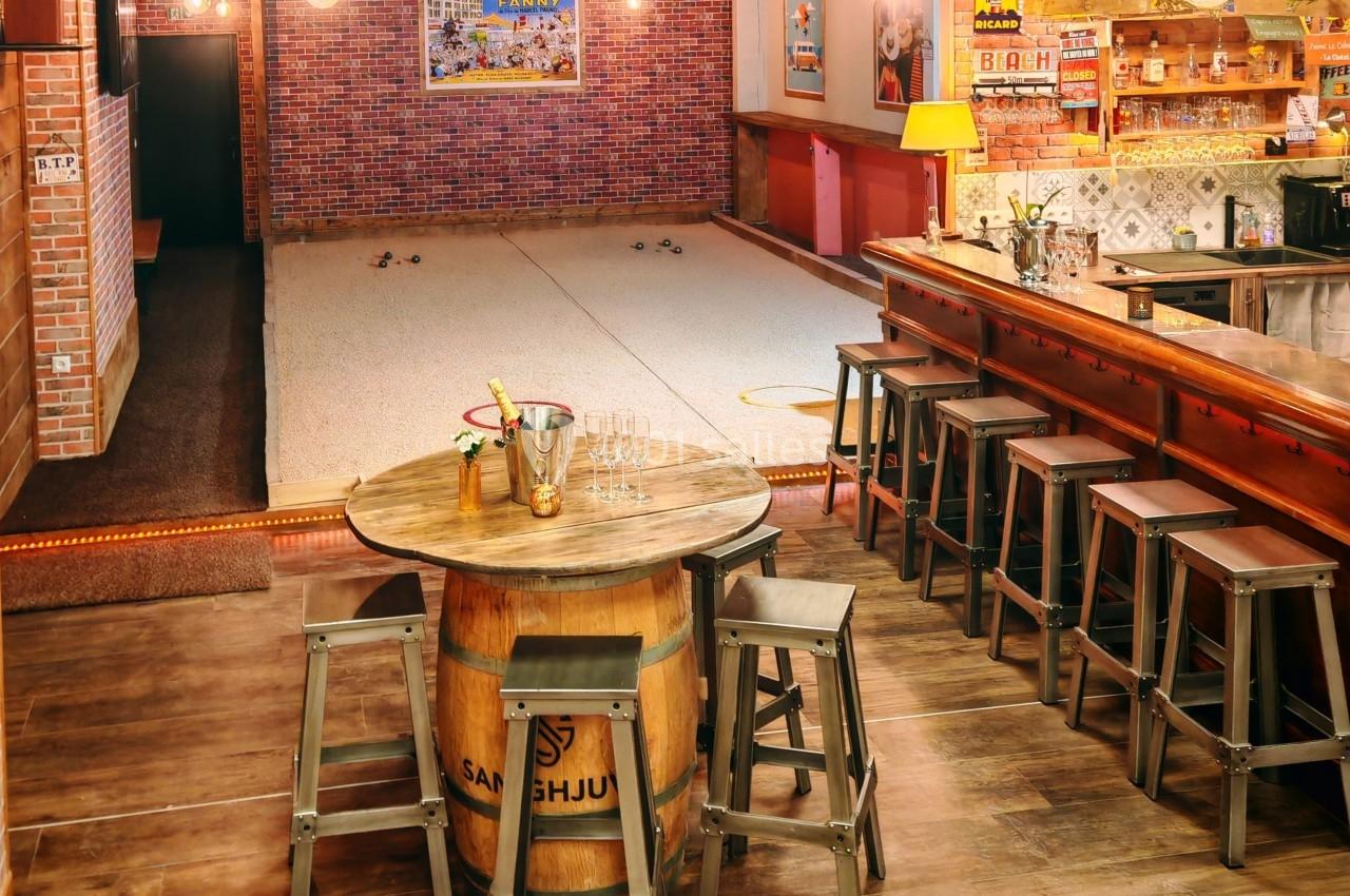 Salle conviviale avec bar en bois, table tonneau, tabourets et espace de jeu intérieur sur sol sablé.