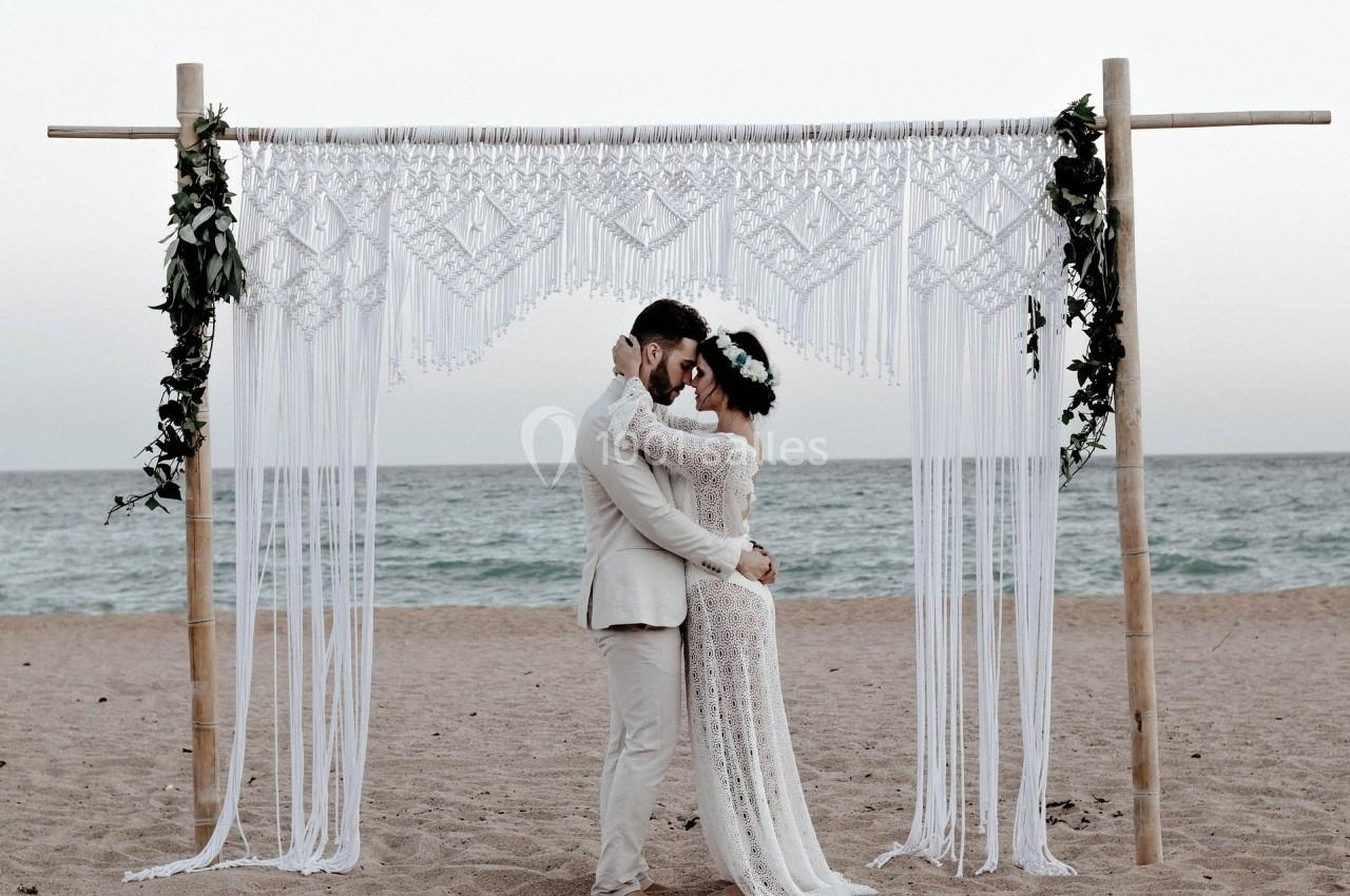 Un couple s'embrasse sous une arche en macramé décorée de feuillage, sur une plage au bord de la mer.