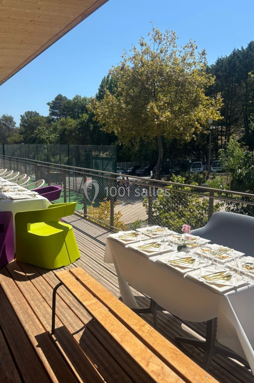 Tables dressées en extérieur sur une terrasse en bois, entourée de chaises colorées et donnant sur un espace arboré.