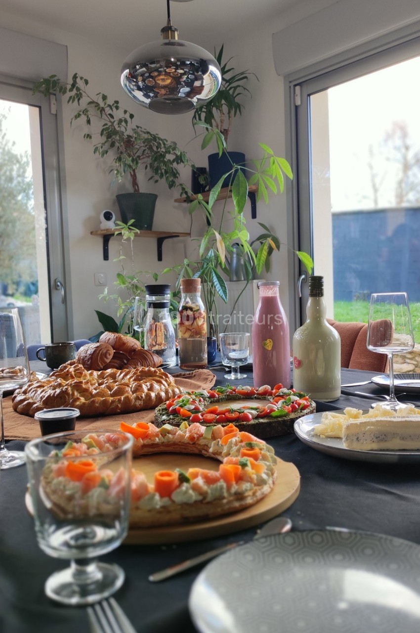 Table dressée avec des tartes salées, viennoiseries, boissons et verres, dans une pièce lumineuse avec vue sur jardin.