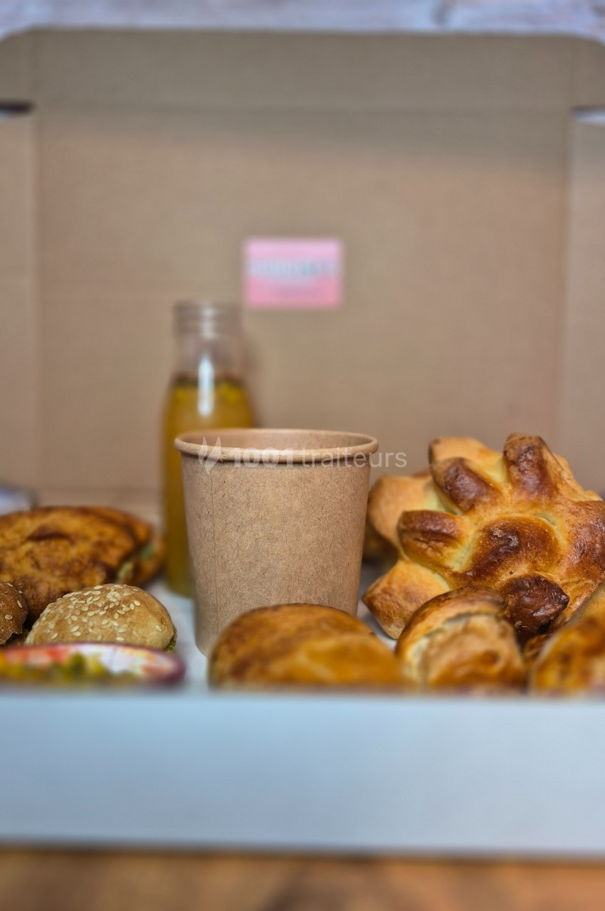 Assortiment de viennoiseries et pain dans une boîte en carton, avec une bouteille de jus et un gobelet en carton.