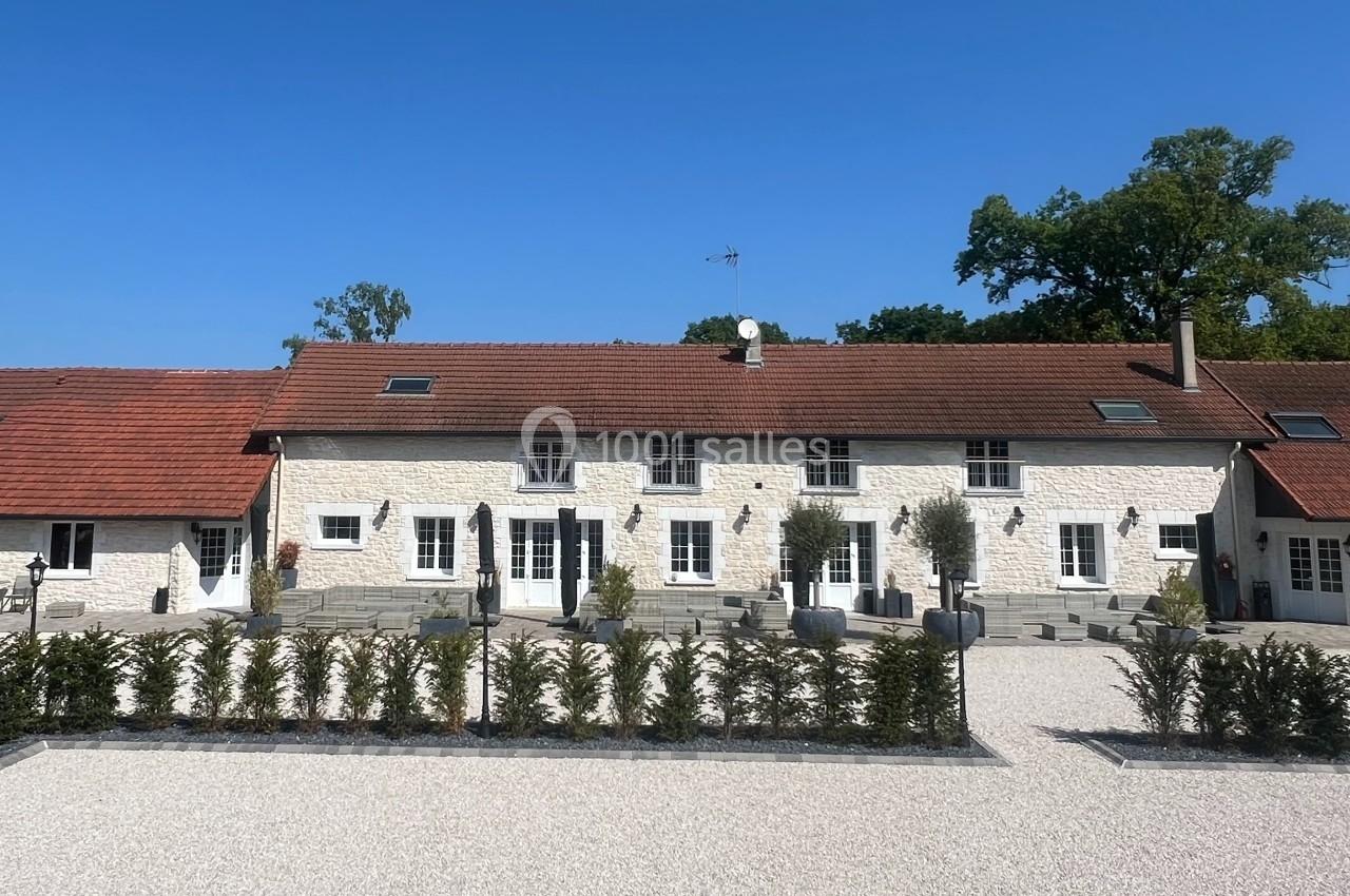 Façade d'une grande maison en pierre avec toit en tuiles rouges, entourée d'une cour gravillonnée et de haies.