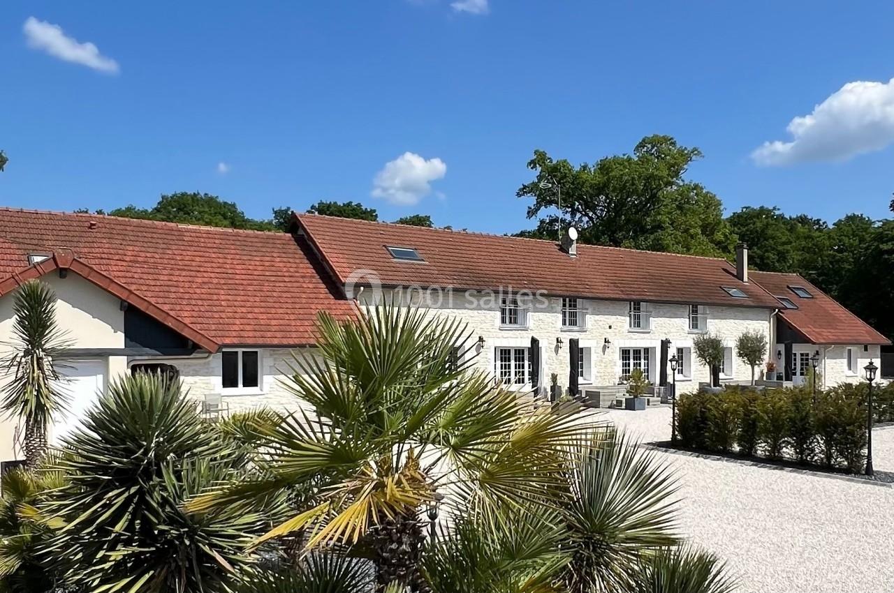 Grande maison en pierre avec toit en tuiles rouges, entourée de végétation et d'une cour gravillonnée sous un ciel bleu.