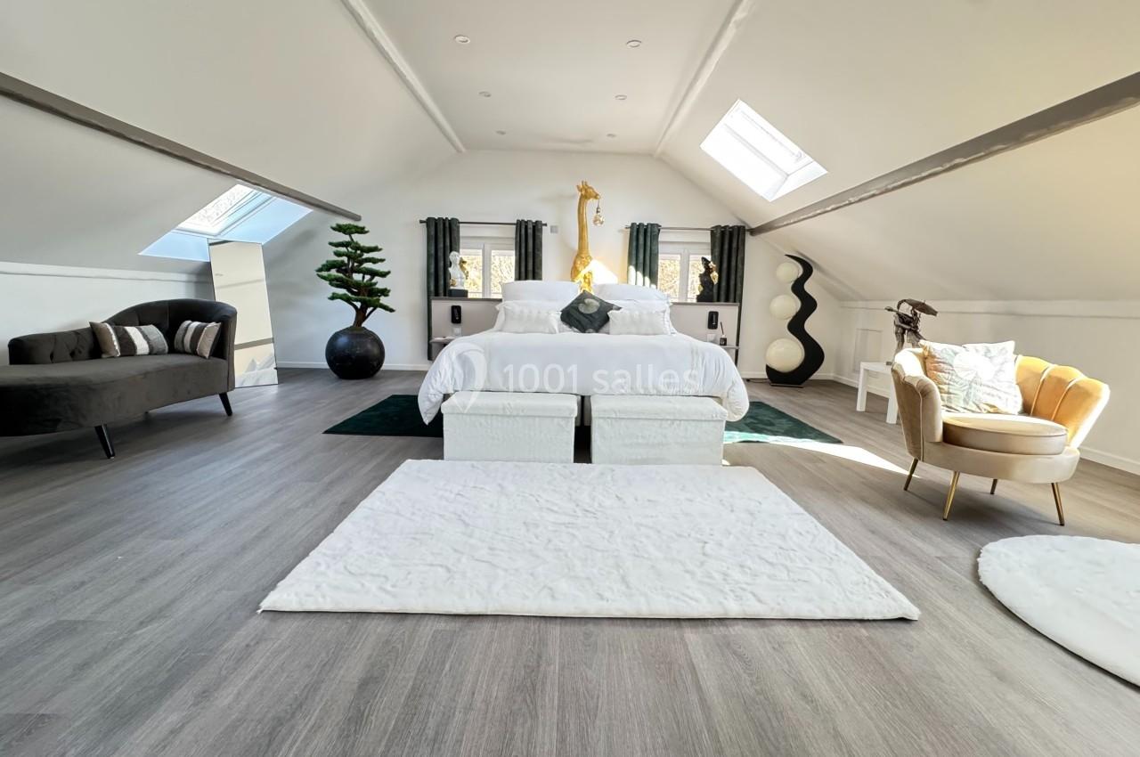 Chambre mansardée lumineuse avec lit central, fauteuils, canapé, décoration moderne et fenêtres de toit.