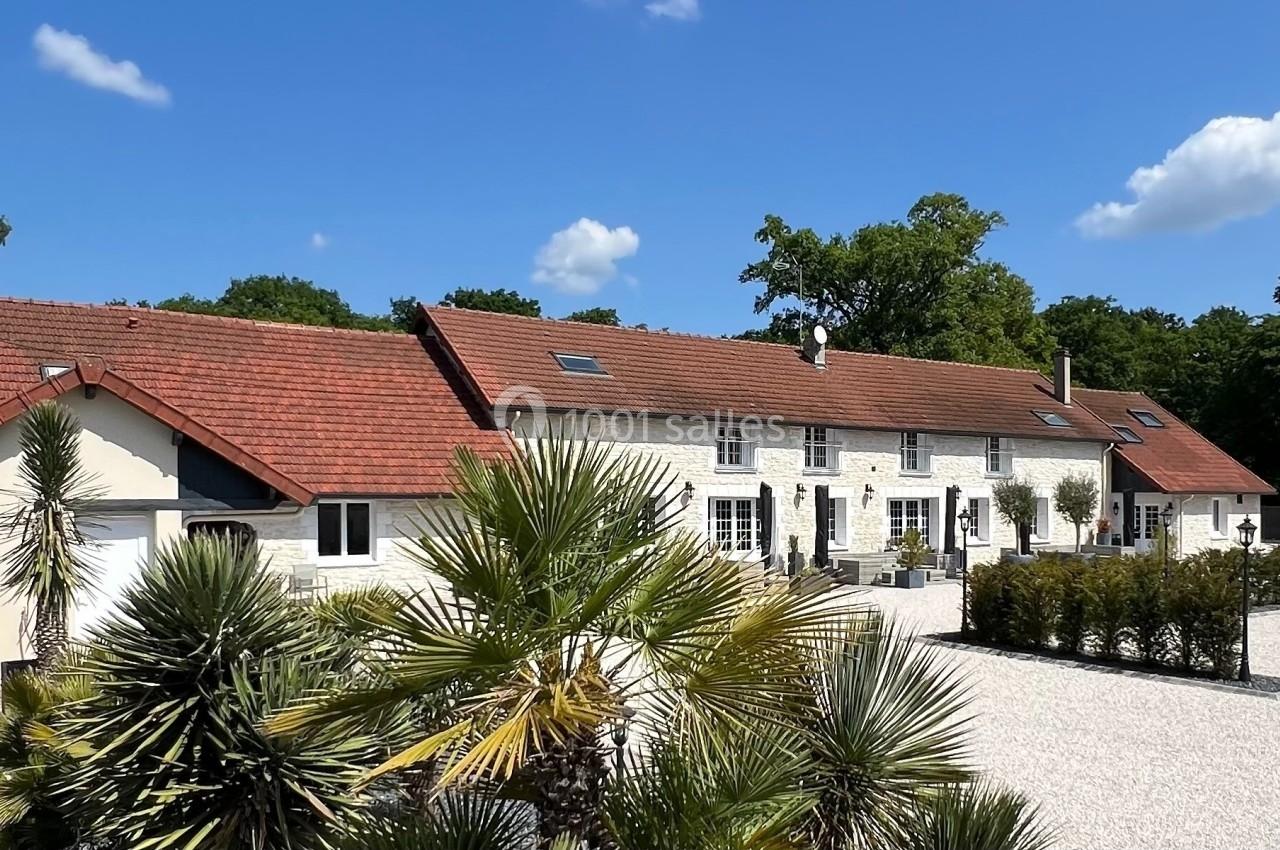 Grande maison en pierre avec toit en tuiles rouges, entourée de végétation et d'une cour gravillonnée sous un ciel bleu.