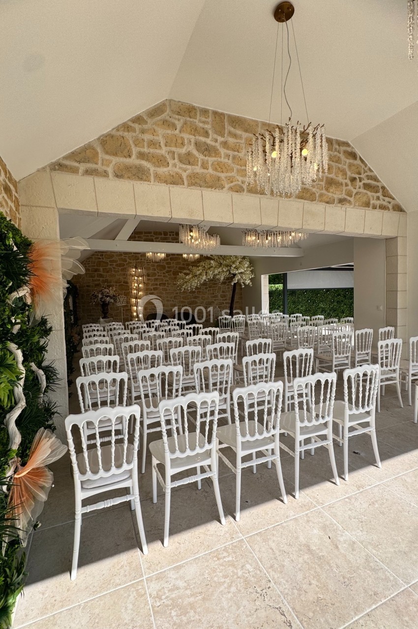 Salle de réception lumineuse avec des chaises blanches alignées, murs en pierre et décorations végétales.