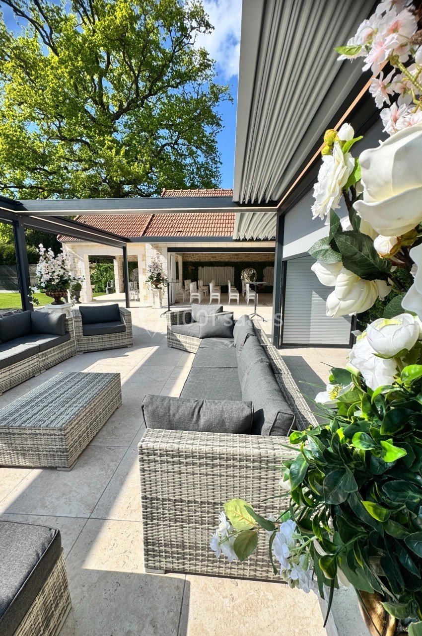 Salon de jardin en rotin gris sous une pergola, entouré de fleurs et donnant sur une terrasse ensoleillée.