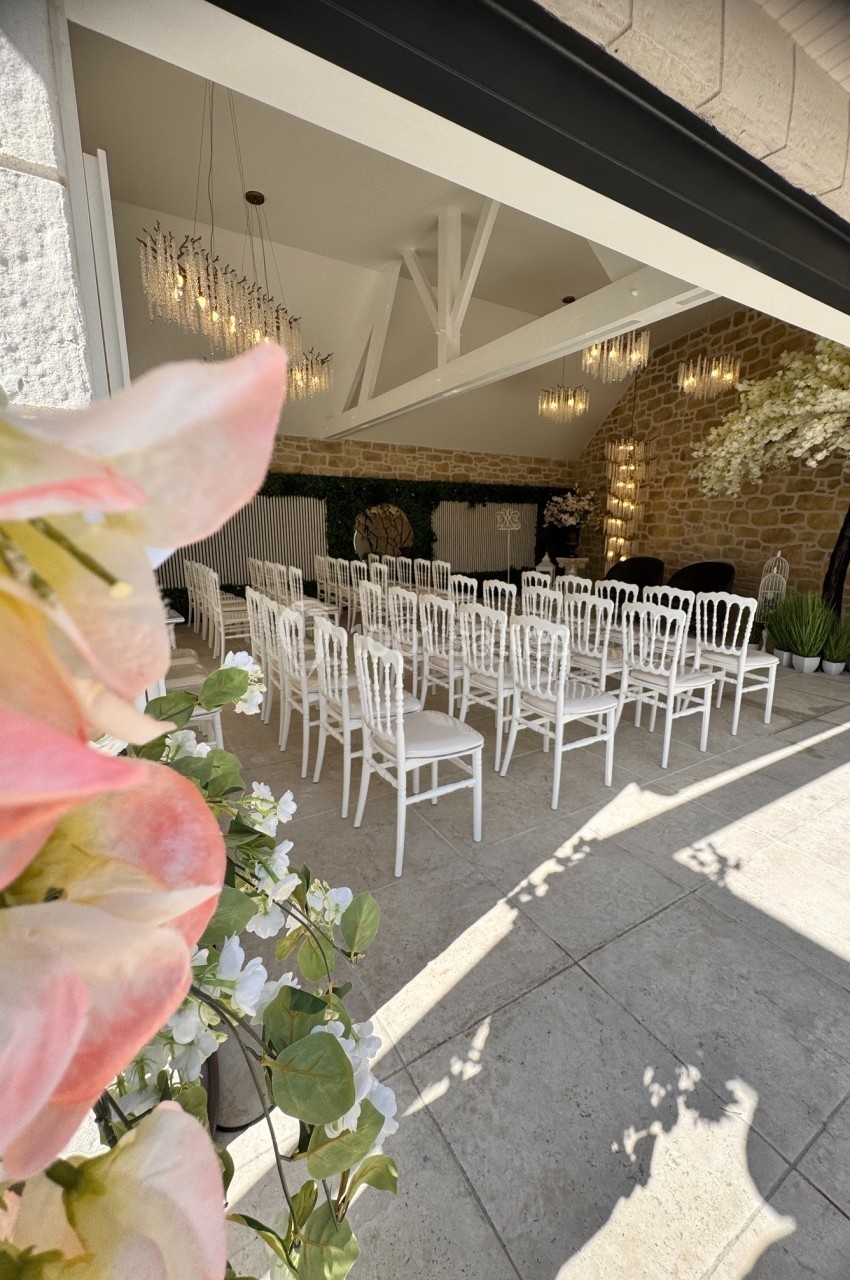 Salle de cérémonie lumineuse avec des chaises blanches alignées, décorée de fleurs et de murs en pierre.