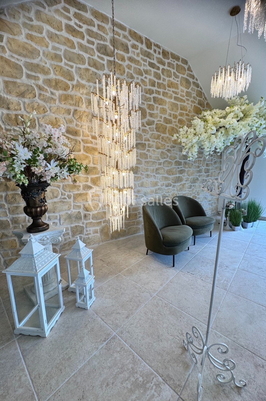 Salle lumineuse avec murs en pierre, lustres en cristal, fauteuils verts, lanternes blanches et décor floral élégant.