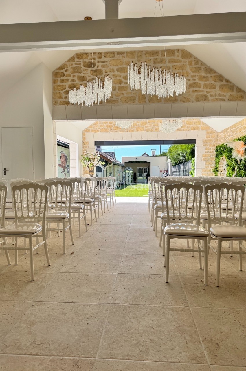 Salle lumineuse avec des chaises blanches alignées, vue sur un jardin à travers une grande ouverture.