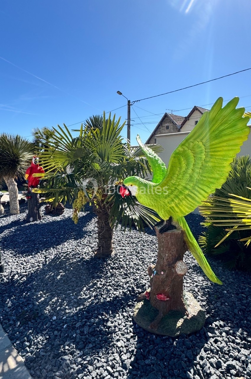 Statues colorées de perroquets sur des troncs d'arbres dans un jardin avec palmiers et ciel dégagé.