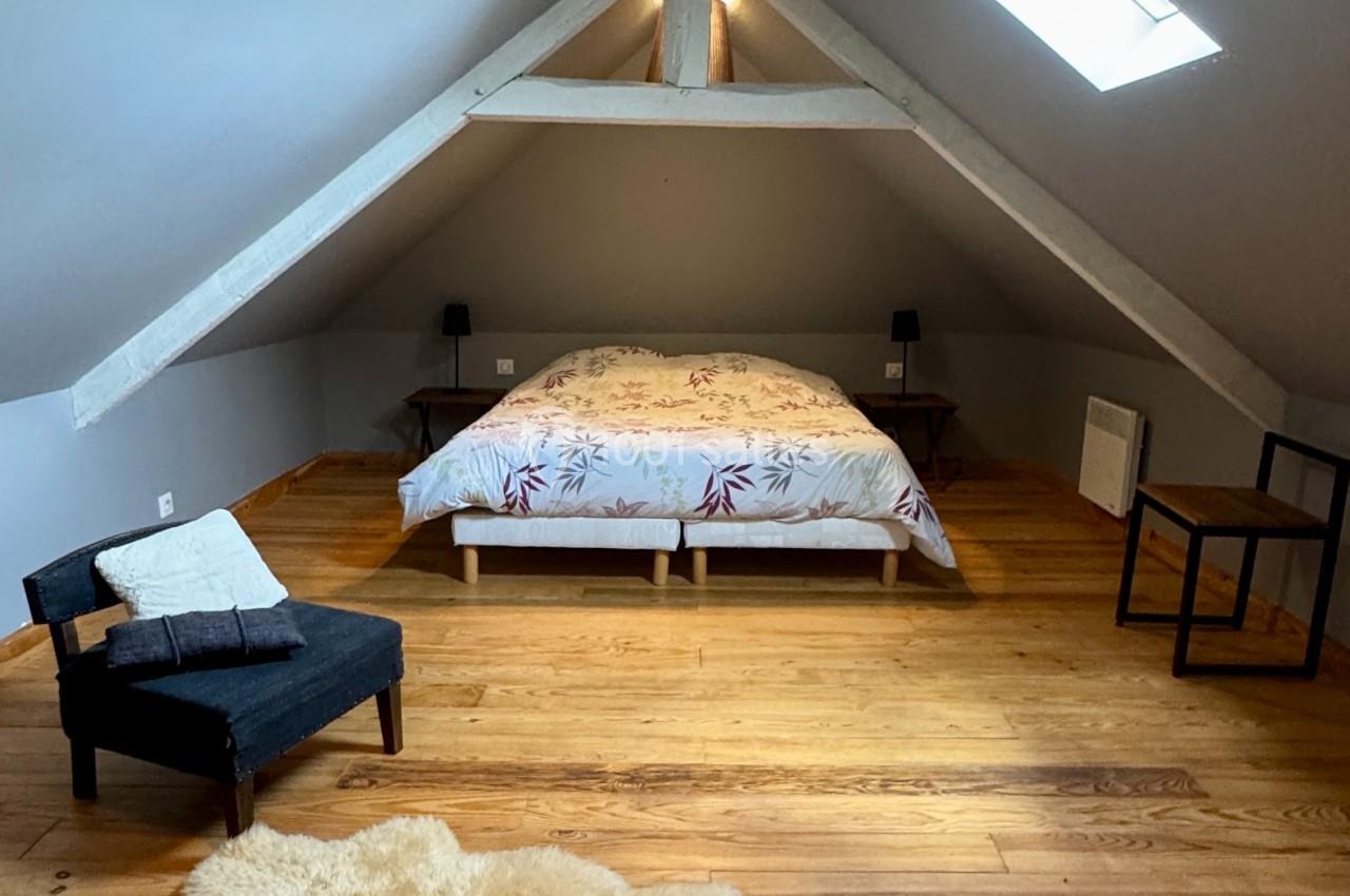 Chambre mansardée avec lit double, parquet en bois, fauteuil, table d'appoint et lumière naturelle provenant d'une fenêtre.