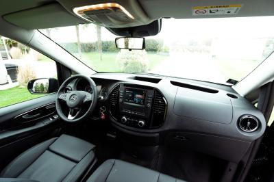 Une femme charge une valise jaune dans le coffre ouvert d'un van noir stationné dans un environnement boisé.