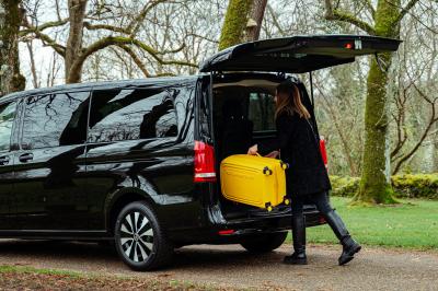 Une femme charge une valise jaune dans le coffre ouvert d'un van noir stationné dans un environnement boisé.