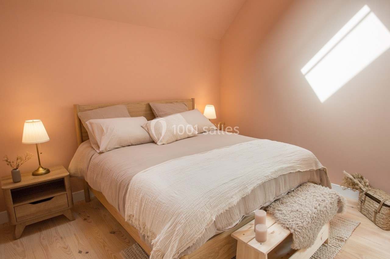 Lit double avec draps beiges dans une chambre aux murs rose pâle, éclairée par une lumière naturelle.
