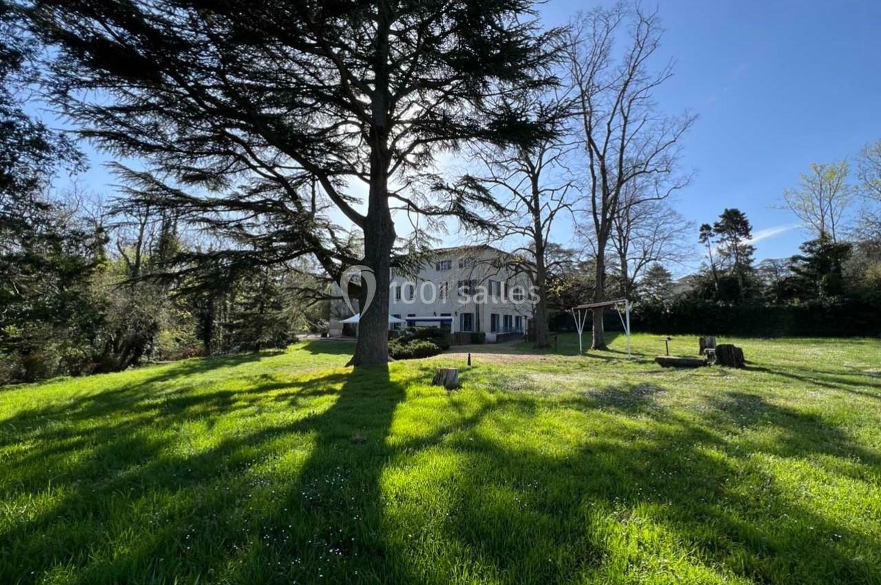 Grande maison blanche entourée d'arbres, avec une balançoire et une pelouse ensoleillée au premier plan.